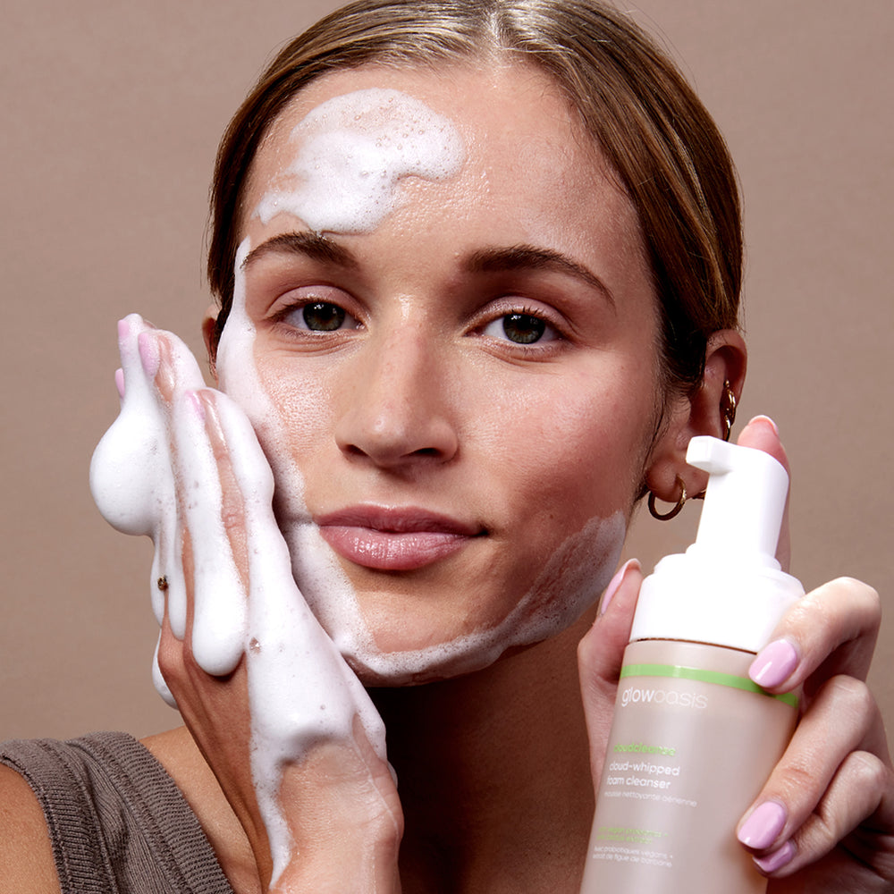 Female model with glowoasis vegan probiotics cloudcleanse cloud whipped gentle daily foam cleanser applied on cheeks and forehead while holding sustainable dispenser in hand.