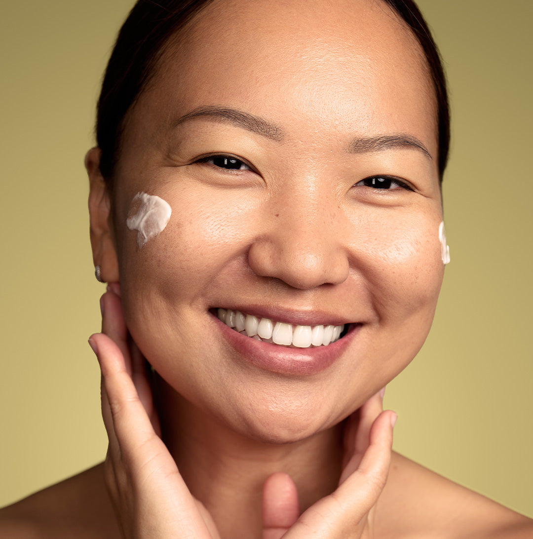 Smiling female with dabs of cream moisturizer on her cheeks, standing in front of a yellow background.