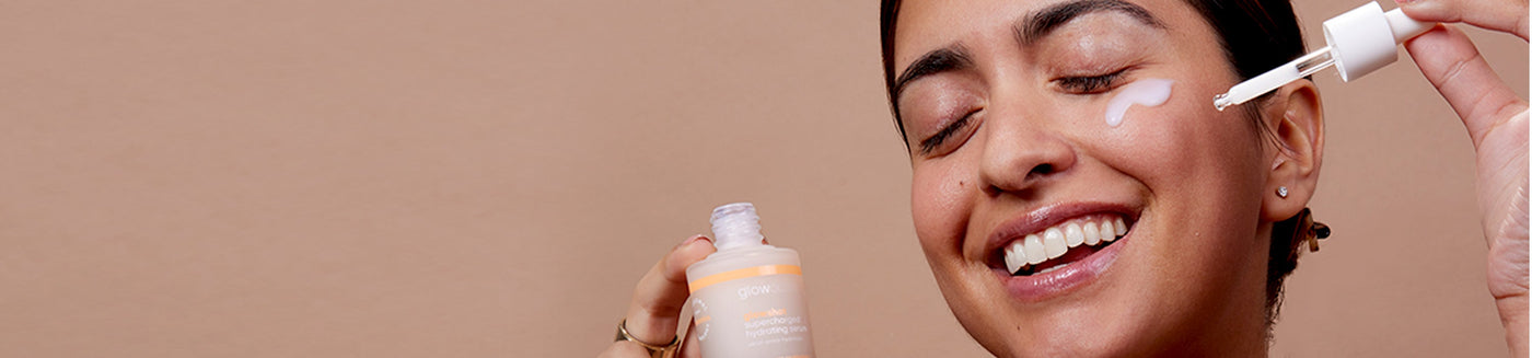 Smiling female model pouring glowoasis vegan probiotics glowshot supercharged hydrating serum with a dropper onto cheek while holding the serum in hands.