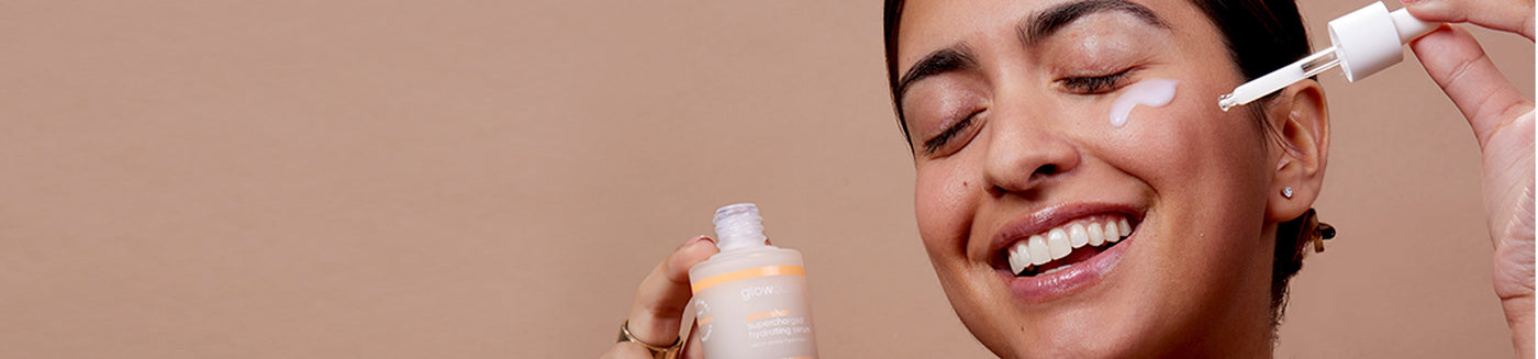 Smiling female model pouring glowoasis vegan probiotics glowshot supercharged hydrating serum with a dropper onto cheek while holding the serum in hands.