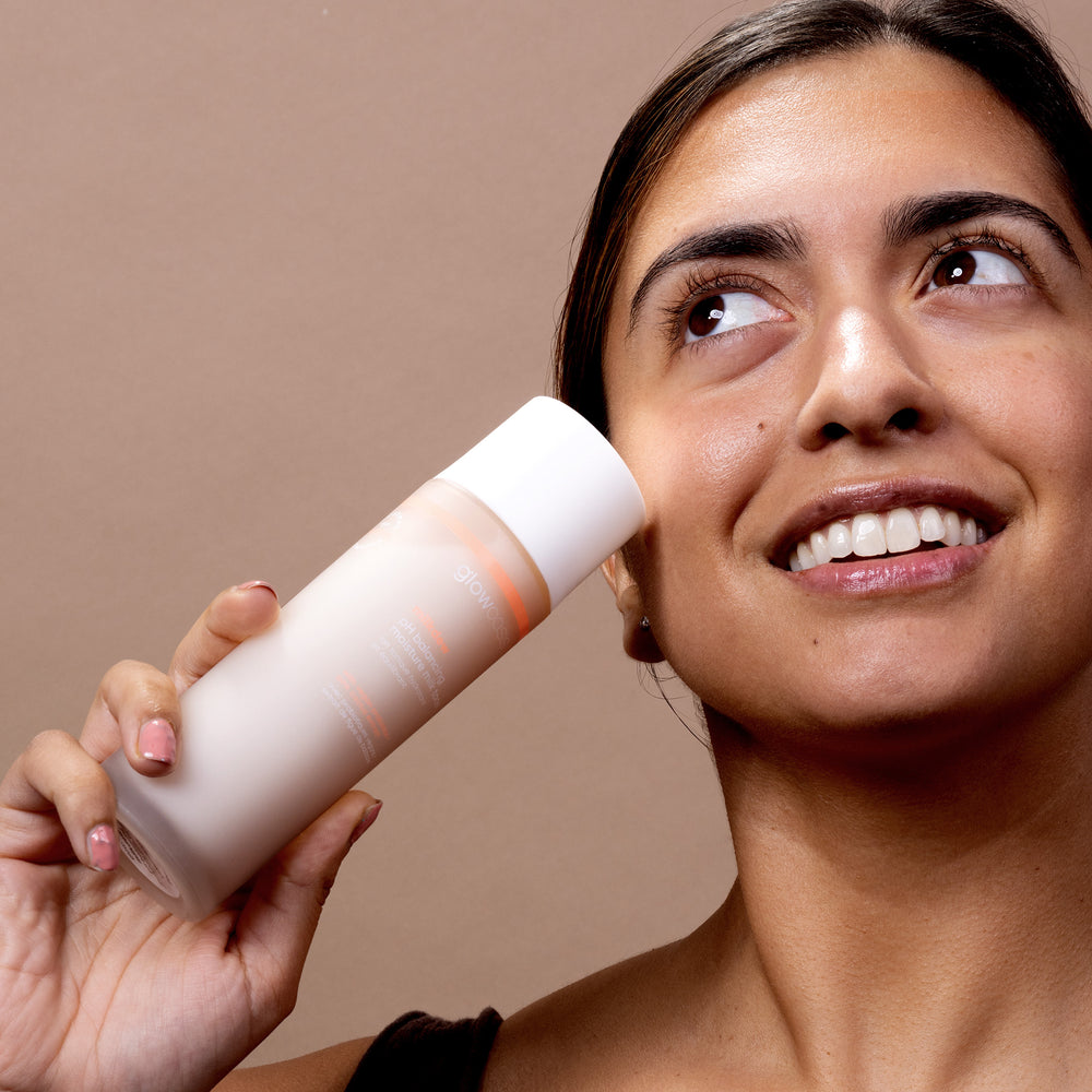 Smiling female model holding glowoasis vegan probiotics milkdew pH balancing moisture milk toner bottle to cheek.