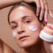 woman holding probiotics + murumuru hydra surge moisturizer with swatches on her cheeks